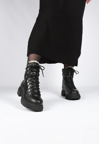 Zwarte leren veterlaarzen met zilverkleurige hardware, dikke rubberen zolen, een gestructureerde afwerking en een gewatteerde enkelsluiting, gecombineerd met een zwart kledingstuk.