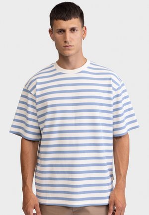 Jeune homme aux cheveux courts et foncés portant une chemise à manches courtes à rayures bleues claires et blanches, debout devant un fond uni.