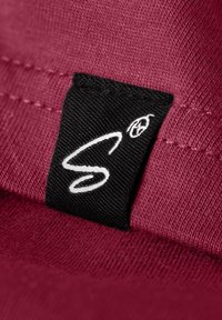 Primo piano di un tessuto viola con un'etichetta nera caratterizzata da cuciture bianche e un logo stilizzato a "S". Texture liscia con dettagli di cucitura visibili.
