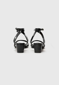 Sandales à talons noires avec une finition brillante, dotées de fines brides croisées et d'une boucle, surélevées par un talon carré.