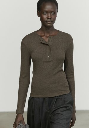 Femme aux cheveux courts et bouclés portant une chemise henley marron à manches longues et un pantalon sombre taille haute, tenant un objet gris texturé dans la main.