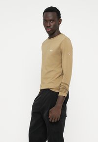 Camicia marrone a maniche lunghe con scollatura tonda e logo ricamato sul petto, abbinata a pantaloni cargo neri con tasche laterali.