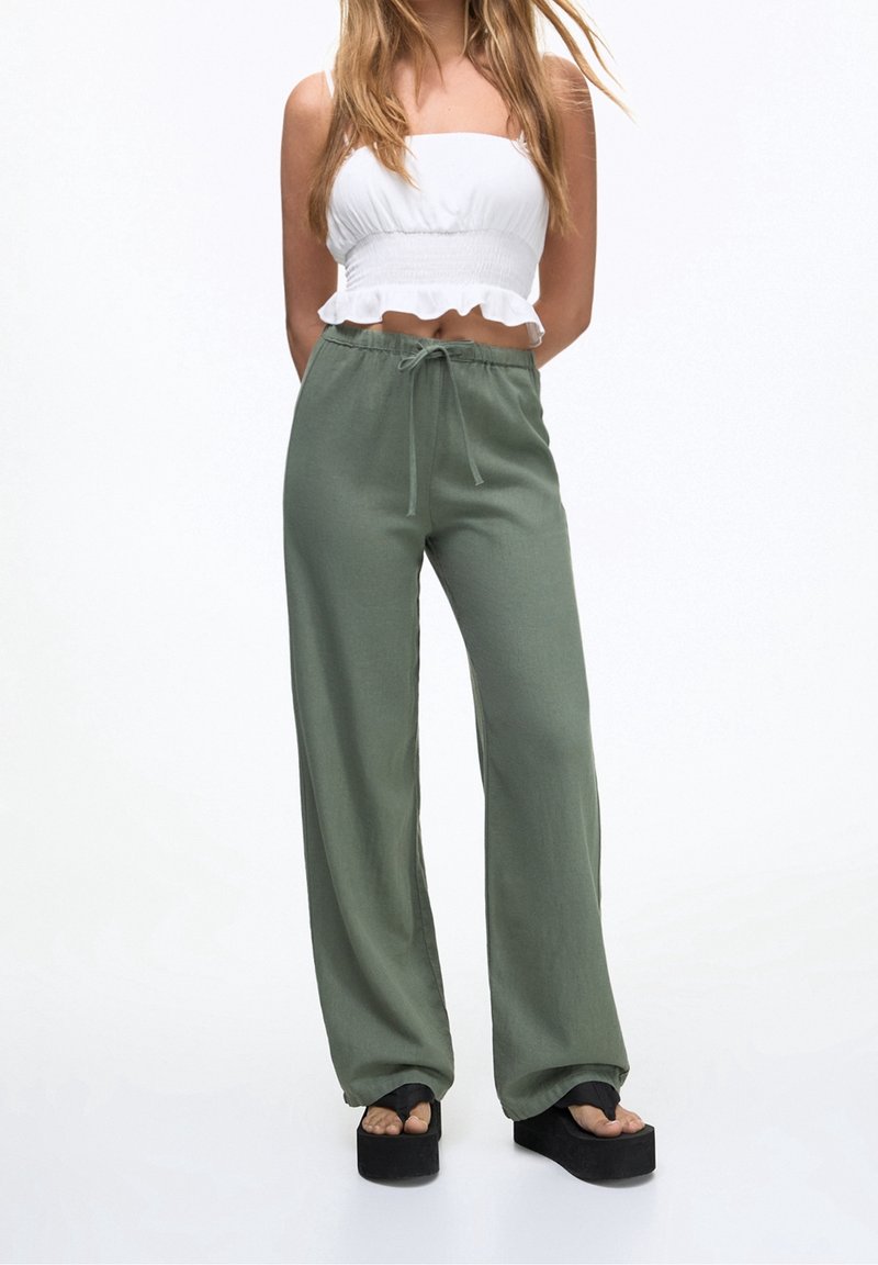 Pantalon ample couleur vert olive en tissu léger, avec une ceinture réglable, associé à un crop top blanc smocké. Sandales noires à plateforme.