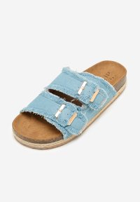 Sandalias deslizantes de denim azul claro con correas deshilachadas, herrajes en tono dorado y una plantilla de ante marrón sobre una suela de yute trenzado.