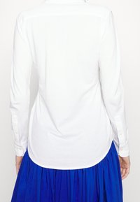 Vit långärmad skjorta med knäppta manschetter, med rundad nederkant och mjuk textur, ihop med en blå, veckad kjol.