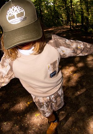 Sweatshirt tan avec des manches camouflage, shorts beiges, et une casquette olive arborant un logo blanc. Des bottes marron complètent l'ensemble.