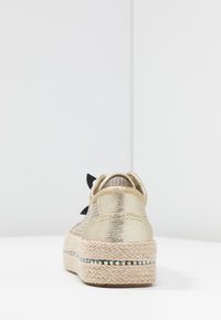 Zapatilla espadrille metálica dorada con detalle de lazo negro, acabado texturizado y suela de yute con una franja decorativa de cuentas.