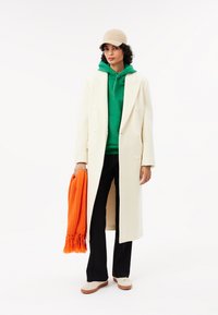 Abrigo de doble botonadura en crema sobre una sudadera verde, pantalones negros y zapatillas blancas, sosteniendo una bufanda de punto naranja. Gorra beige complementa el atuendo.