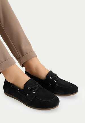 Person trägt schwarze Wildleder-Schnürslipper mit braunen Sohlen und hochgekrempelte beige Hosen vor weißem Hintergrund.