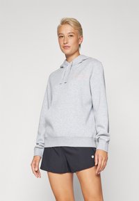 Felpa grigia chiara con tasca a marsupio, cappuccio con lacci e polsini a coste. Presenta un piccolo logo rosa sul petto. Indossata con pantaloni corti neri.