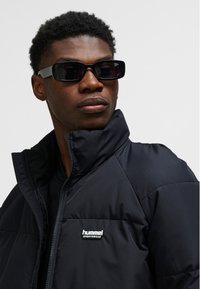 Sort dunjakke med høj krave og lynlås, med en lille hvid logo på brystet. Modellen bærer rektangulære sorte solbriller.