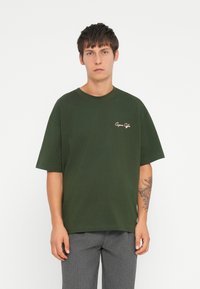 Camiseta de algodón verde oscura con mangas cortas y cuello redondo. Presenta un pequeño logotipo bordado en el pecho de color contrastante.