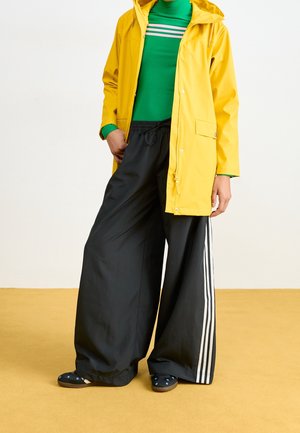 Personne portant un imperméable jaune, une chemise à rayures vertes, un pantalon large noir avec des bandes blanches sur les côtés, et des chaussures noires avec des motifs bleus, debout sur un sol jaune.