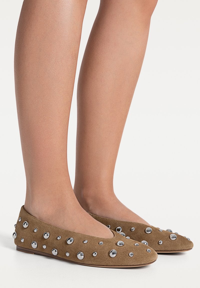 Jambes portant des ballerines en daim marron ornées de clous argentés brillants de différentes tailles sur un fond blanc.