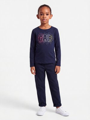 Enfant debout portant un t-shirt bleu marine à manches longues avec un texte coloré "GAP", un pantalon bleu marine et des baskets blanches sur fond blanc.