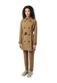 Brun trenchcoat med bælte, dobbeltbrystet design, sorte knapper, parret med matchende brune bukser og lyse sneakers.
