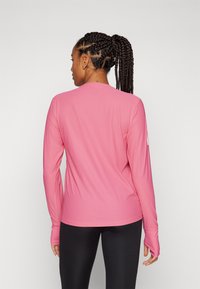 Maglietta sportiva rosa a maniche lunghe con una superficie liscia. Presenta fori per il pollice sulle maniche e un semplice scollo a girocollo. Cuciture piatte per il comfort.