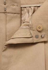 Tissu beige à texture lisse, doté d'une fermeture éclair et d'un bouton, accompagné de pièces métalliques argentées et de détails de couture subtils.