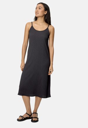 Mujer con un vestido midi negro sin mangas, con tirantes finos, y sandalias negras planas, de pie contra un fondo claro y liso.