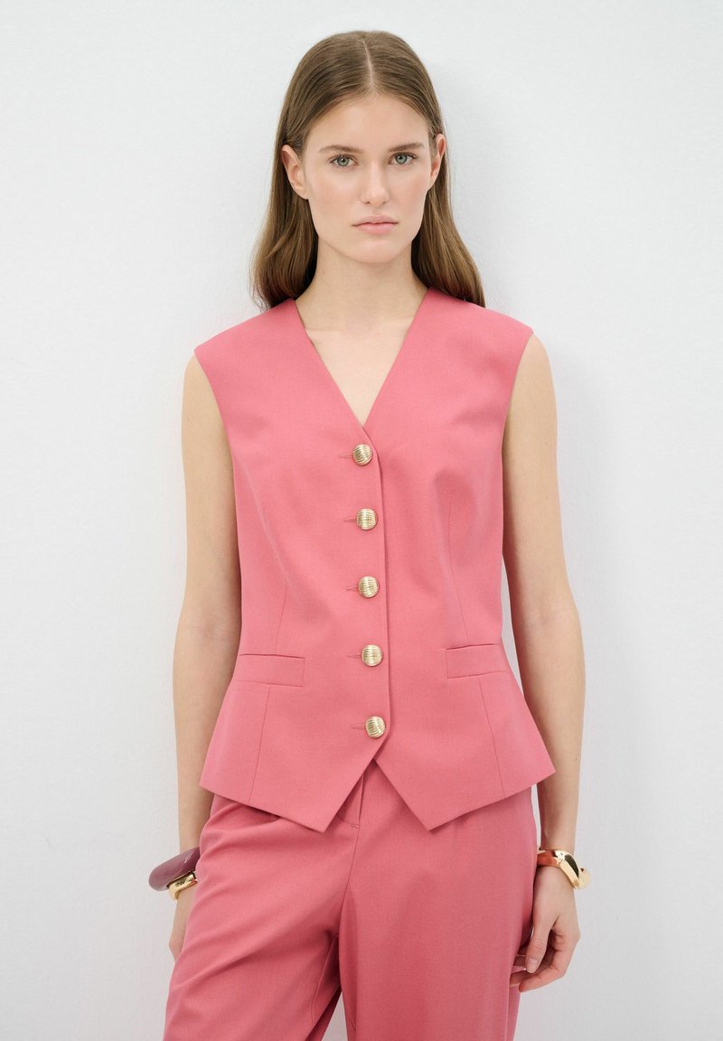 Gilet sans manches rose avec un col en V, comportant cinq boutons dorés, une coupe ajustée et des poches latérales. Porté avec un pantalon assorti.