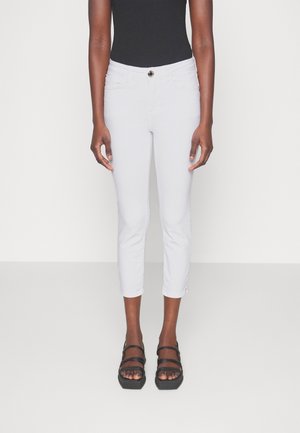 Personne portant un pantalon blanc court ajusté, un haut noir sans manches et des sandales à plateforme noires à bouts ouverts sur un fond uni.