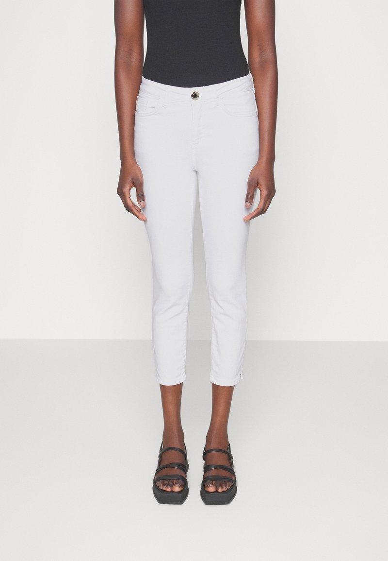 Personne portant un pantalon blanc court ajusté, un haut noir sans manches et des sandales à plateforme noires à bouts ouverts sur un fond uni.