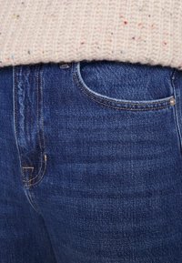 Granatowe dżinsy w klasycznym kroju, z subtelnym wzorem teksturowym i kontrastowymi stębnowaniami, częściowo przykryte jasnym swetrem z dzianiny.