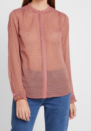Vrouw draagt een roze blouse met lange mouwen, een klein geometrisch patroon en knoopmanchetten, gecombineerd met een blauwe spijkerbroek, en staat tegen een effen achtergrond.
