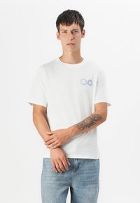 Hvit bomull T-skjorte med korte ermer, med blå smilefjes og krysse-symboler på venstre bryst. Modellen har på seg lyseblå jeans.
