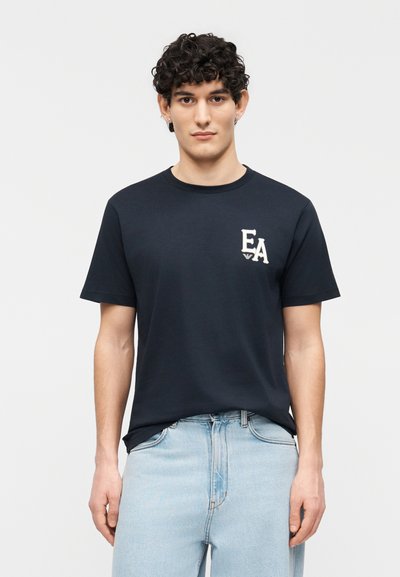 Νέος άνδρας με κατσαρά μαλλιά που φοράει μαύρο μπλουζάκι με το λογότυπο EA και ανοιχτόχρωμο τζιν, στέκεται μπροστά σε ένα απλό λευκό φόντο.