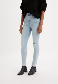 Jeans ajustados de color azul claro con un acabado desgastado y un ligero desgarro en la rodilla, combinados con botines negros y un suéter negro texturizado.