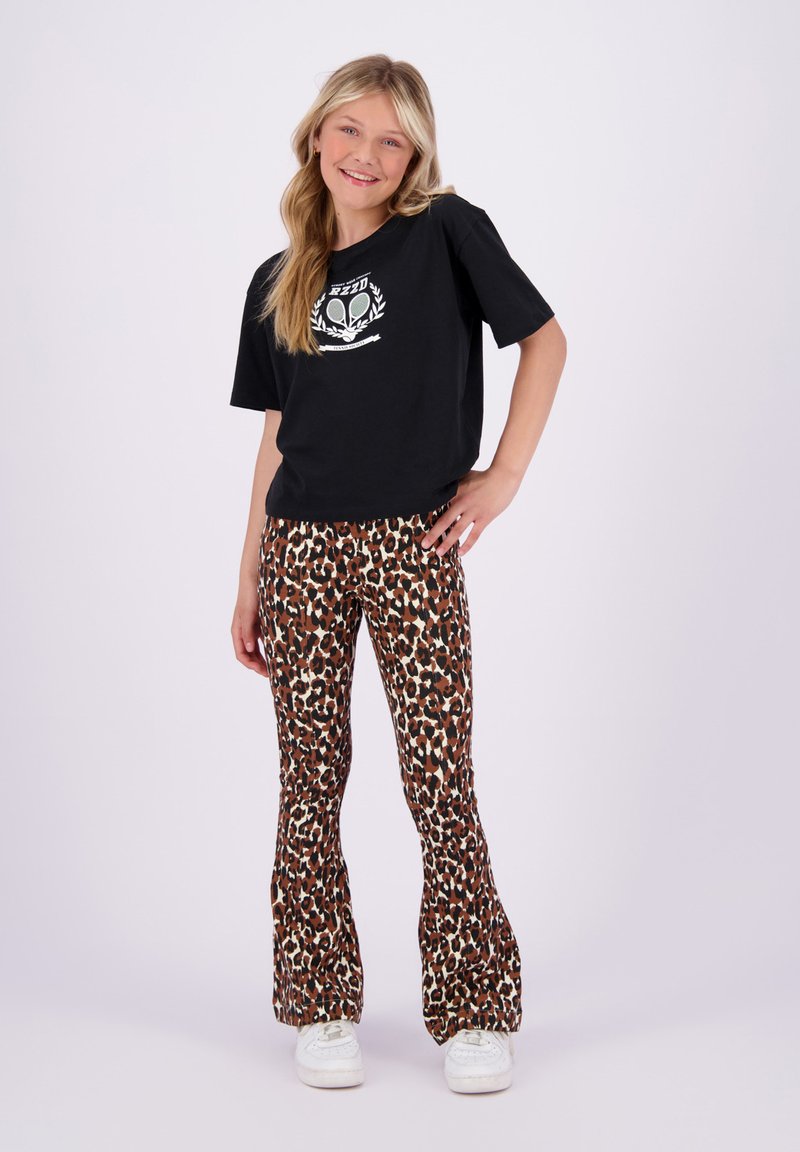 Svart kortermet t-skjorte med et alienmotiv. Brune flare leggings med leopardmønster. Hvite sneakers. Enkel bakgrunn.