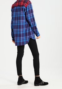 Camicia a quadri blu e rossa con un retro più lungo, abbinata a pantaloni skinny neri e stivaletti con dettagli in metallo.