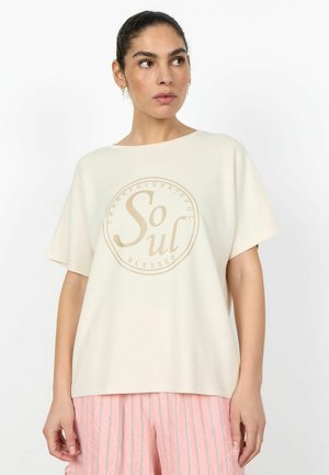 Kvinde iført cremefarvet oversized T-shirt med teksten "Soul" og lyserøde stribede bukser, stående foran en hvid baggrund.