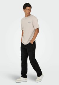 Camiseta de manga corta beige con un gráfico azul, combinada con jeans negros y zapatillas blancas. El modelo está de pie con las manos en los bolsillos.