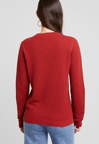 Pull rouge tricoté avec un motif texturé, col rond, poignets et ourlet côtelés. Porté avec un jean bleu clair.