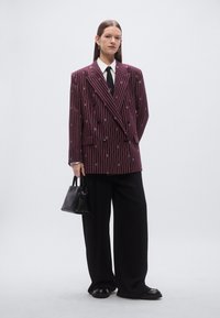 Dvoredni, bordo srajčni blazer s širokimi ovratniki, kombiniran z belo srajco, črno kravato, širokimi črnimi hlačami in majhno črno torbico.