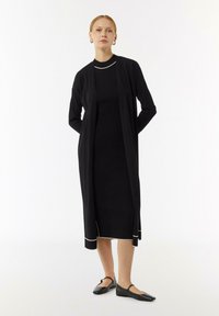 Schwarzes Kleid mit gerippter Textur, das mit einem langen Cardigan kombiniert ist. Beide Teile haben einen weißen Rand. Flache schwarze Schuhe runden das Outfit ab.