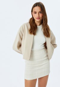 Beige cropped jack met zachte stof, lange mouwen en een rits aan de voorkant, gecombineerd met een gestructureerde off-white minirok.