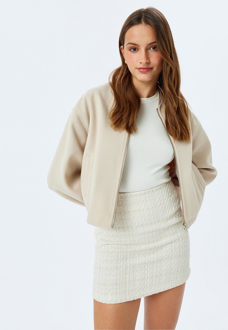 Beige cropped jack met zachte stof, lange mouwen en een rits aan de voorkant, gecombineerd met een gestructureerde off-white minirok.