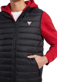 Chaleco acolchado negro con cierre de cremallera, que lleva una sudadera con capucha roja debajo. Incluye un parche con el logo y bolsillos laterales.