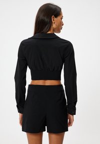 Camicetta nera corta con maniche lunghe e colletto, caratterizzata da un dettaglio arricciato sul retro. Abbinata a pantaloni corti neri a vita alta.