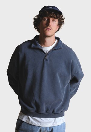 Námornícky modrý fleece pulóver so zipsovým golierom, spustenými ramenami, rebrovanými manžetami a uvoľneným strihom, nosený cez biele tričko.