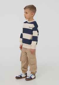 Bambino che indossa una felpa a righe blu navy e crema con la scritta "Divertiti", pantaloni cargo beige e sneaker blu e bianche.