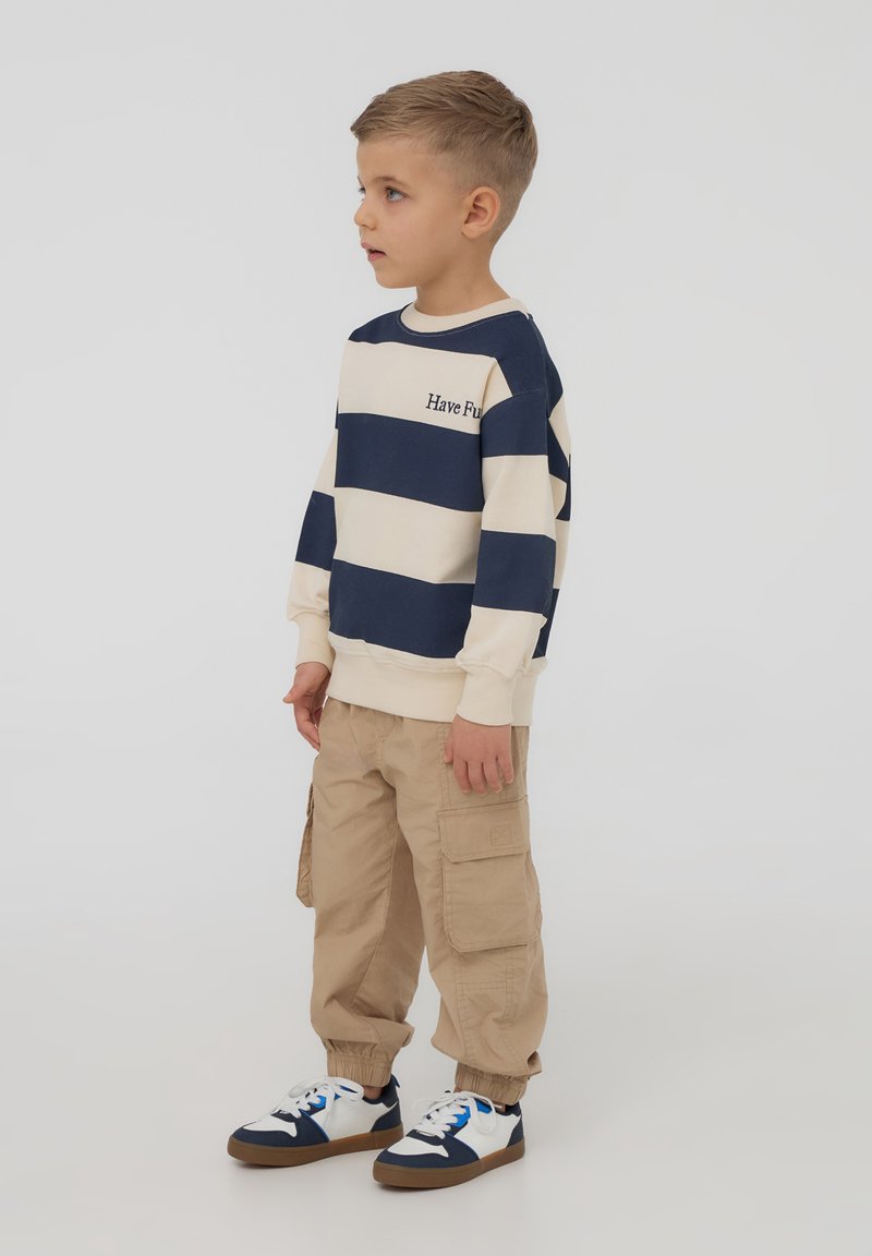 Bambino che indossa una felpa a righe blu navy e crema con la scritta "Divertiti", pantaloni cargo beige e sneaker blu e bianche.