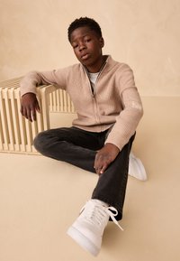 Cardigan beige à fermeture éclair avec texture côtelée, porté sur une chemise grise. Jean noir et baskets blanches complètent la tenue. Assis sur un fond pâle.