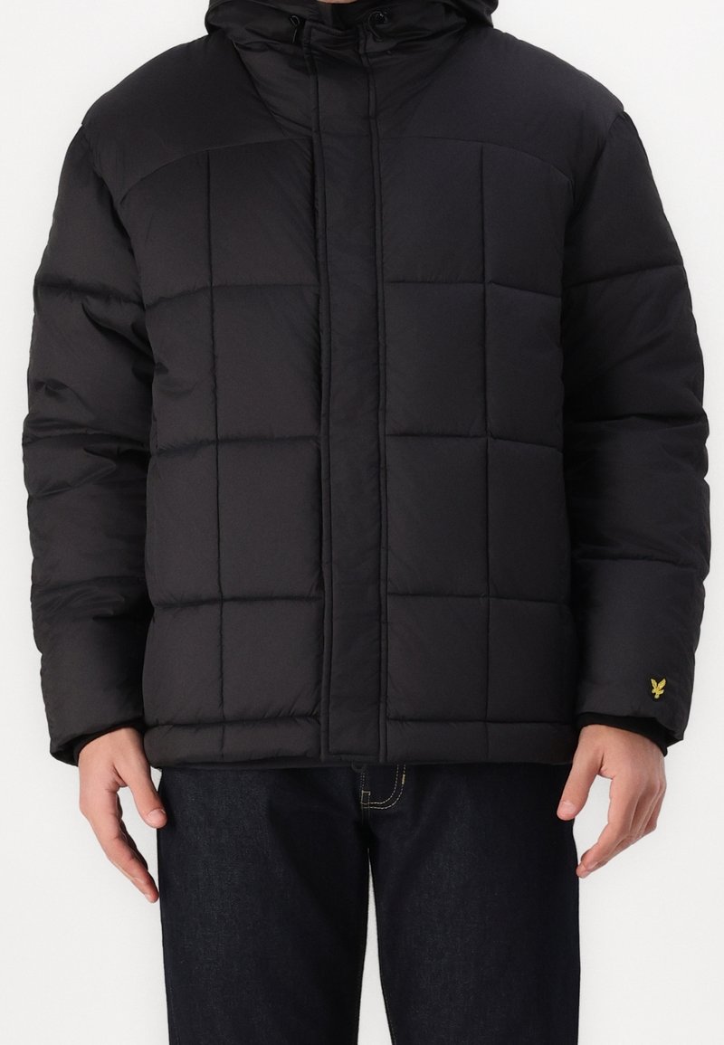 Veste matelassée noire avec un design matelassé, une capuche et une fermeture éclair à l'avant. Présente un petit logo jaune sur la manche gauche. Le tissu semble lisse.