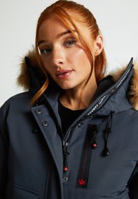 Tumši pelēks parka mētelis ar kažokādas odere jaku, rāvējslēdzēja un pogu aizdari, ar sarkaniem akcentiem un sarežģītām izšūšanas detaļām.