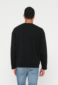 Άνδρας που φαίνεται από πίσω, φοράει απλό μαύρο φούτερ με μακριά μανίκια και ανοιχτόχρωμο μπλε τζιν, μπροστά σε απλό λευκό φόντο.