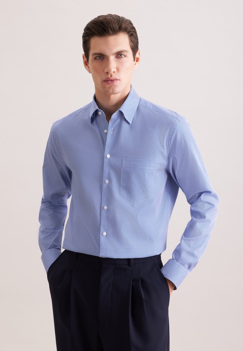 Jeune homme portant une chemise à manches longues bleu clair à motifs, rentrée dans un pantalon plissé bleu foncé, debout avec les mains dans les poches.